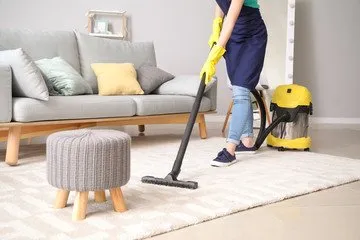 A person in blue attire and yellow gloves is vacuuming a rug in a living room. The room has a gray sofa with colorful pillows, a small wooden stool, and a yellow vacuum cleaner. The focus is on the cleaning task and the cozy living room setting.