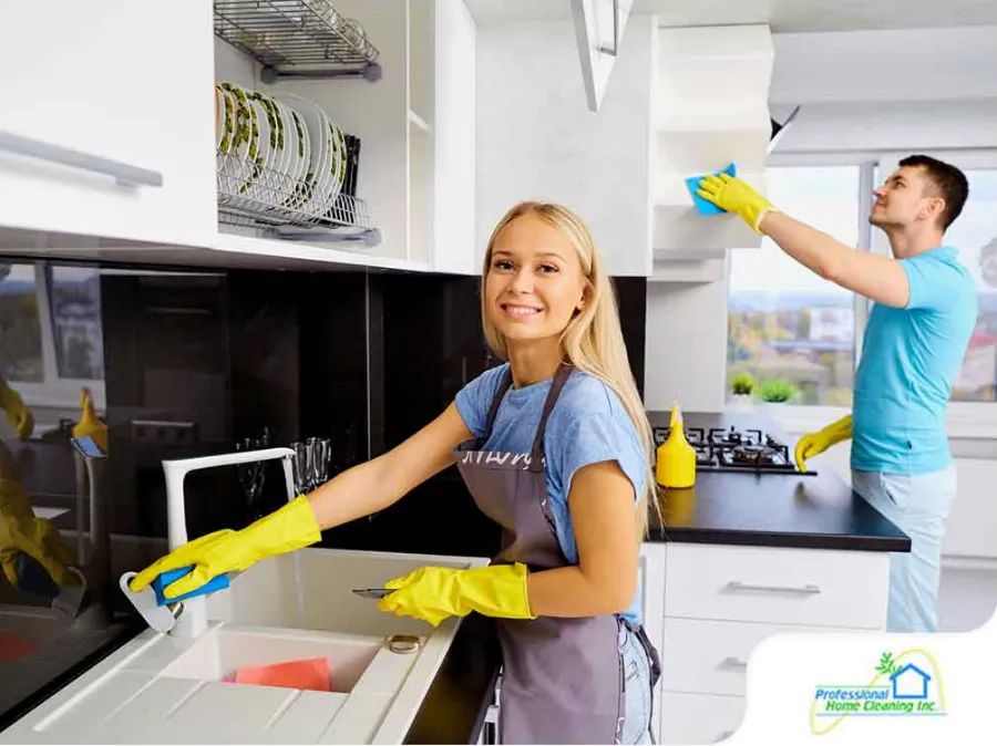 A smiling woman in a blue t-shirt and yellow cleaning gloves is wiping a white kitchen countertop. In the background, a man in a blue t-shirt and yellow gloves is cleaning the cabinets. The kitchen is modern with white cabinets and black countertops. A logo for "Professional Home Cleaning Inc." is visible in the corner, suggesting a promotional or advertising image.