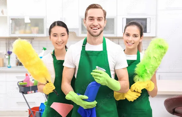 Three people wearing green aprons and yellow gloves are smiling at the camera, holding cleaning supplies. The man in the center is holding a blue spray bottle, while the women on either side are holding yellow and green feather dusters.