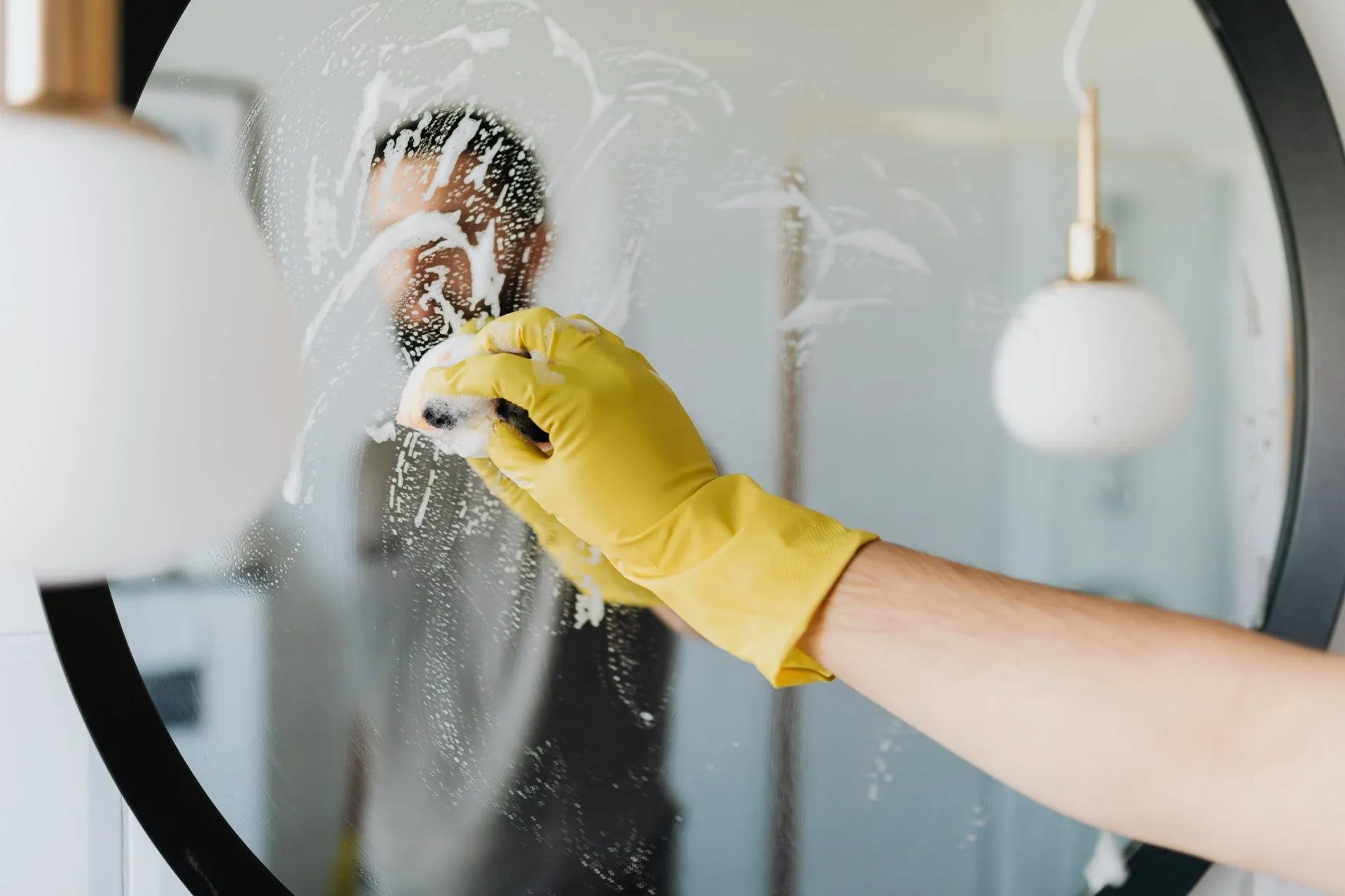 A person's hand in a yellow cleaning glove is wiping soapy suds on a mirror, reflecting their blurred image, with a pendant light visible in the background.