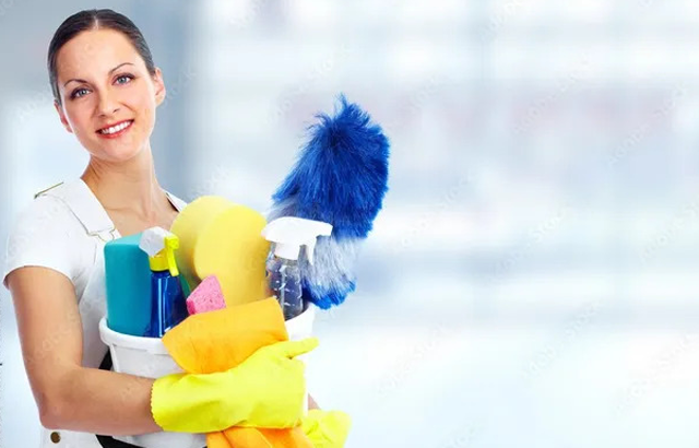 A smiling woman wearing yellow gloves is holding a bucket filled with cleaning supplies, including spray bottles and sponges. A blue feather duster stands out among the supplies.